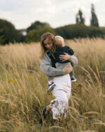 Mama przechodzi przez łąkę, przytula synka siedzącego w szarym nosidełku wspomagającym na biodro DereDere; widoczny miękki naramiennik, wysokie trawy i wieczorne światło.