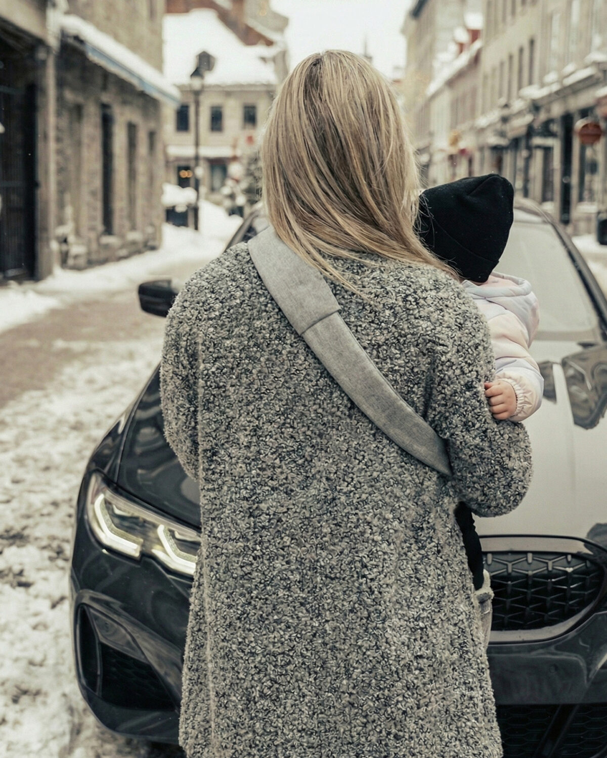Widok od tyłu: mama z dzieckiem na biodrze, szeroki miękki naramiennik nosidełka DereDere biegnie przez plecy; zimowa ulica i zaparkowany samochód.