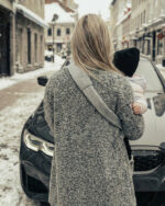 Widok od tyłu: mama z dzieckiem na biodrze, szeroki miękki naramiennik nosidełka DereDere biegnie przez plecy; zimowa ulica i zaparkowany samochód.