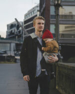 Tata na miejskim moście niesie śpiące dziecko w szarym nosidełku na biodro DereDere; czarna szelka z naramiennikiem.