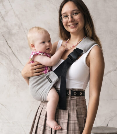 Mama trzyma dziecko w jasnoszarym nosidełku DereDere i uśmiecha się do zdjęcia.