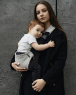 Kobieta w czarnym płaszczu przytula dziecko siedzące w szarym nosidełku ergonomicznym DereDere; widoczna szeroka czarna szelka z klamrą, neutralne, betonowe tło.