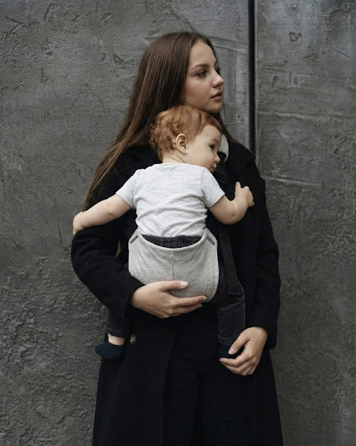 Kobieta w czarnym płaszczu ma nosidełko ergonomiczne i trzyma dziecko oparte o jej ramię; widać szare siedzisko nosidełka DereDere i pas biegnący w dół, proste, szare tło.