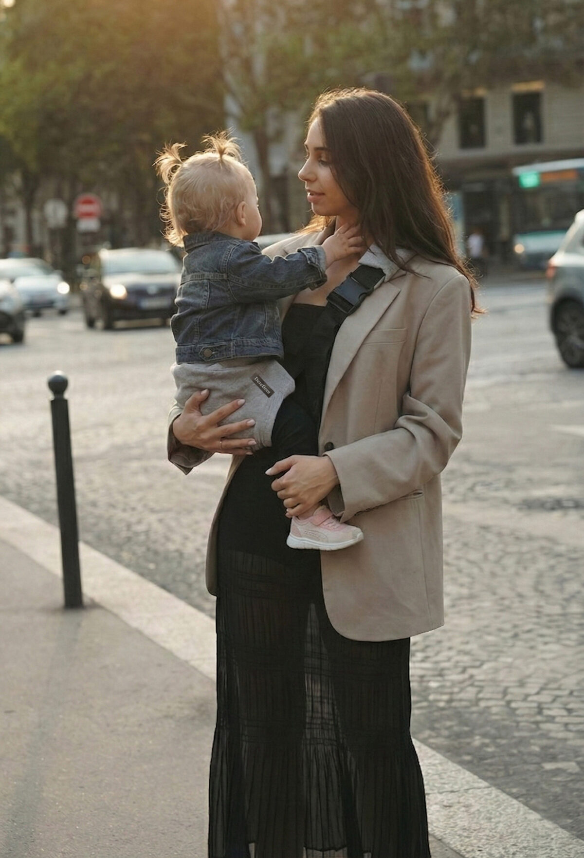 Mama na miejskim spacerze trzyma dziecko w szarym nosidełku na biodro DereDere; czarna szelka z klamrą, złota godzina, ruch uliczny w tle.