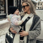 Kobieta w grubym swetrze i płaszczu, w okularach przeciwsłonecznych, trzyma uśmiechnięte dziecko w czapce i pastelowej kurtce; maluch siedzi w szarym nosidełku biodrowym DereDere z czarną szelką i dużą klamrą.