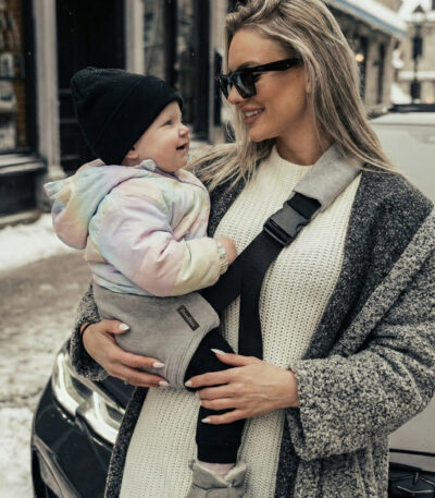 Kobieta w grubym swetrze i płaszczu, w okularach przeciwsłonecznych, trzyma uśmiechnięte dziecko w czapce i pastelowej kurtce; maluch siedzi w szarym nosidełku biodrowym DereDere z czarną szelką i dużą klamrą.