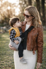 Kobieta w brązowej skórzanej kurtce i okularach słonecznych niesie chłopca w żółtej kamizelce. Dziecko siedzi stabilnie w szarym nosidełku dla dzieci DereDere z szeroką, czarną szelką i miękkim naramiennikiem; tło to zielony park w popołudniowym słońcu.