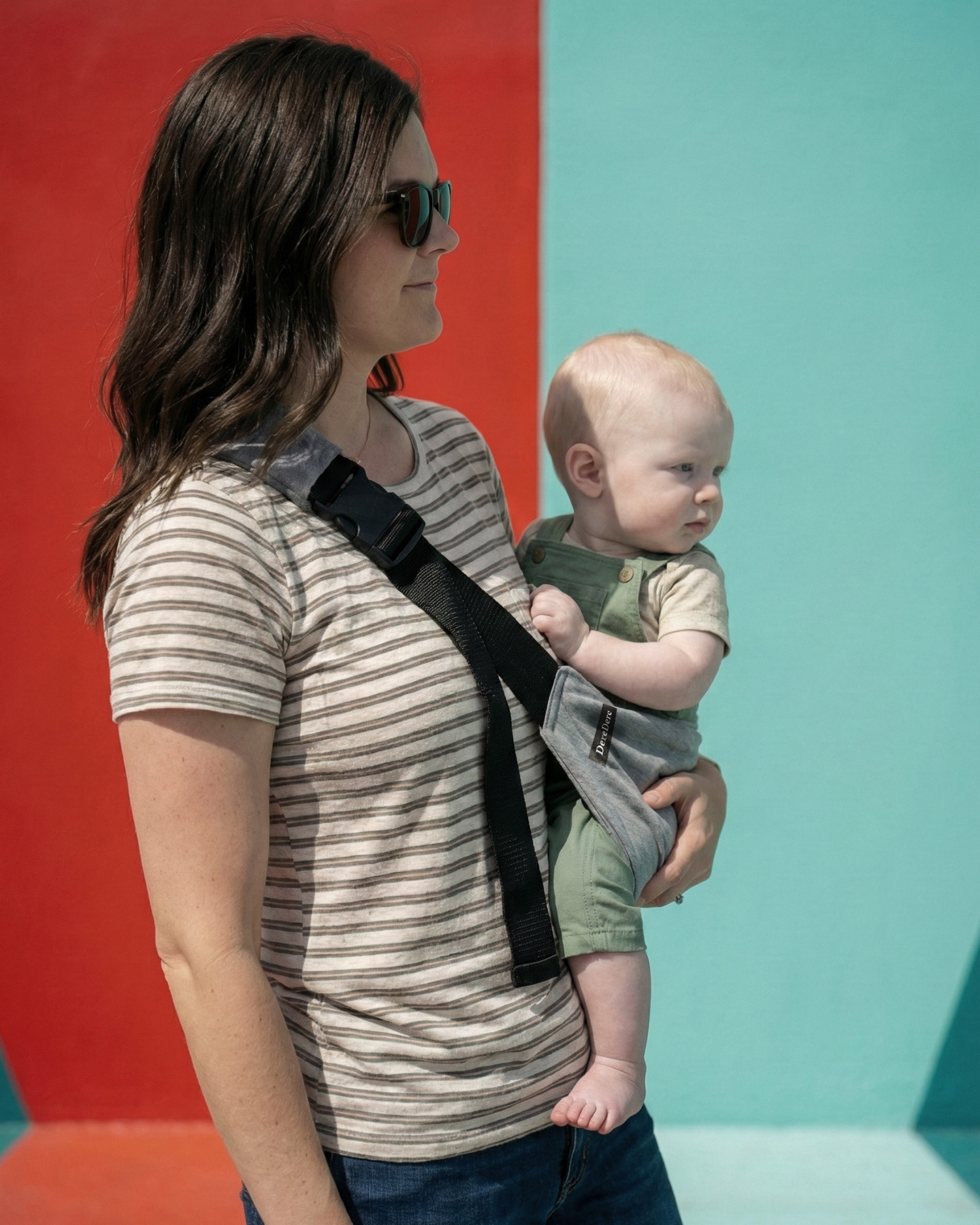 Kobieta w pasiistej koszulce stoi przy geometrycznej, czerwono-turkusowej ścianie i trzyma malucha siedzącego w szarym nosidełku wspomagającym DereDere z szeroką czarną szelką i miękkim naramiennikiem.