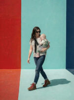 Kobieta w pasiistej koszulce i okularach słonecznych idzie obok czerwono-turkusowej ściany. Dziecko siedzi stabilnie w szarym nosidełku na biodro DereDere z szeroką czarną szelką; casualowy, letni styl.