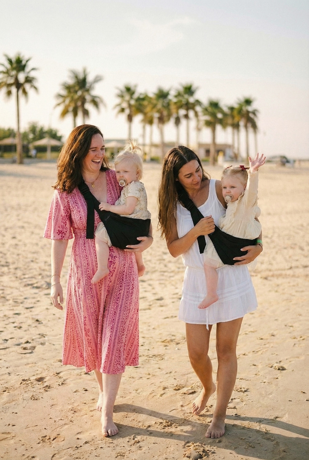 Dwie mamy idą boso po plaży i niosą dzieci w czarnych nosidełkach na biodro DereDere; słoneczne popołudnie, palmy w tle.