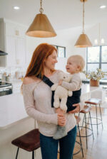 Mama w jasnej kuchni trzyma malucha z pluszowym misiem w czarnym nosidełku wspomagającym DereDere; szeroka szelka z klamrą.