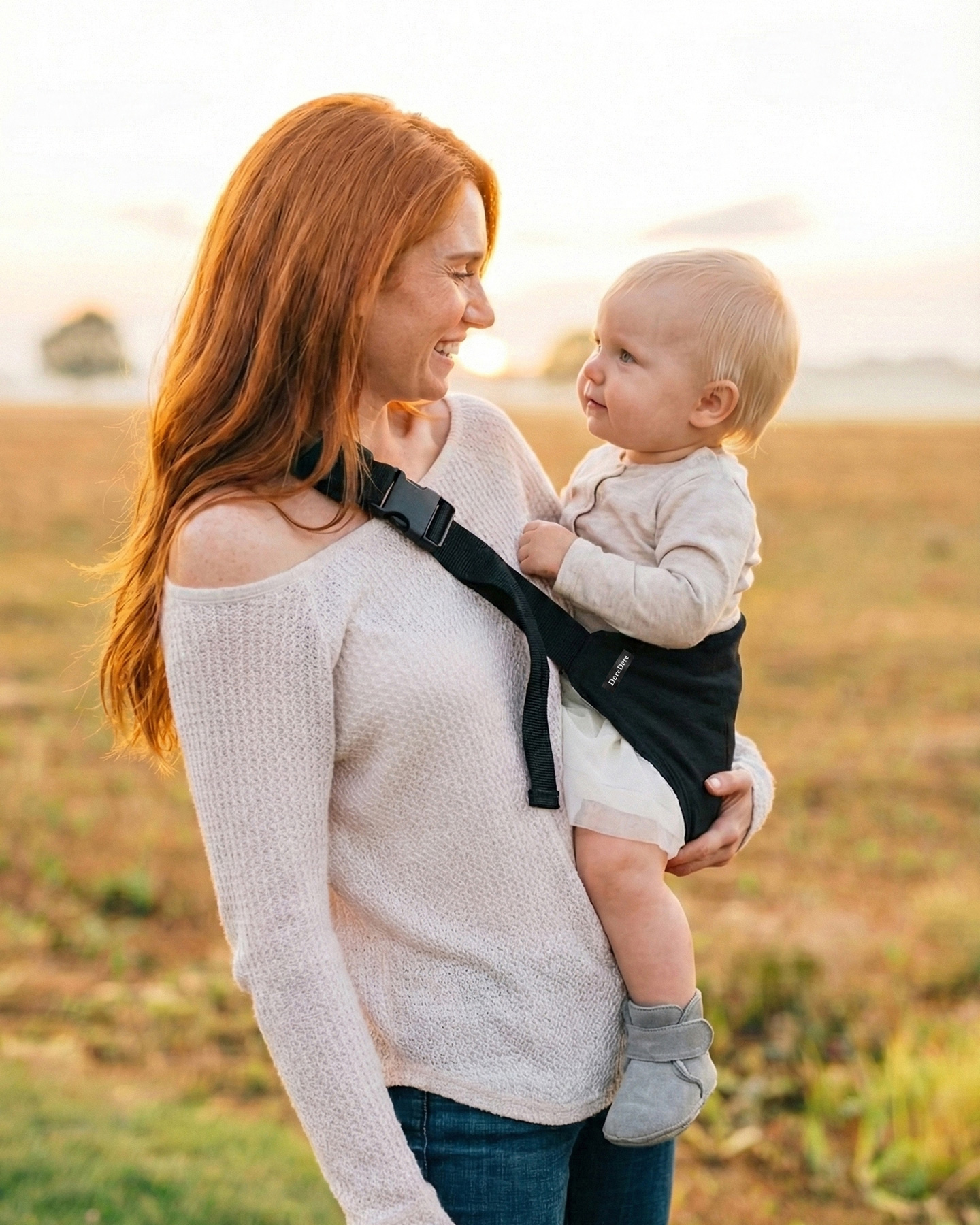 Rudowłosa mama na otwartej łące uśmiecha się do dziecka siedzącego w czarnym nosidełku dla dzieci DereDere; złota godzina, subtelne tło krajobrazu.