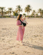 Mama w różowej sukience spaceruje po plaży, ma nosidełko dla dziecka w kolorze czarnym i niesie w nim dziecko; palmy i piasek w tle.
