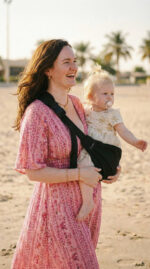 Zbliżenie: uśmiechnięta mama w różowej sukience trzyma córkę w czarnym nosidełku na biodro DereDere; dziewczynka z smoczkiem wskazuje ręką.