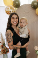 Uśmiechnięta mama trzyma dziecko w czarnym nosidełku DereDere podczas rodzinnej uroczystości.