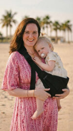 Portret: mama w różowej sukience patrzy w obiektyw, dziecko wtulone w czarne nosidełko dla dzieci DereDere; palmy i plaża w tle.