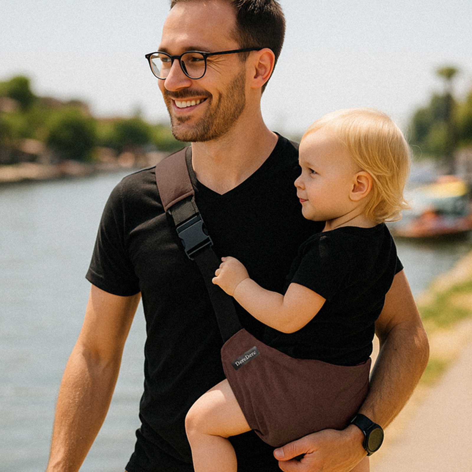 Mężczyzna w czarnej koszulce i okularach idzie bulwarem nad wodą, niosąc dziecko w brązowym nosidełku biodrowym DereDere z czarną szelką i dużą klamrą; maluch siedzi bokiem, patrzy przed siebie.