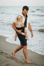 Tata niesie dziecko w czarnym nosidełku dla dziecka DereDere podczas spaceru po plaży.