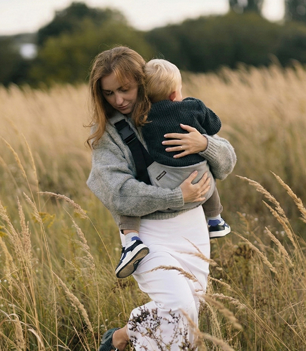 Mama przechodzi przez łąkę, przytula synka siedzącego w szarym nosidełku wspomagającym na biodro DereDere; widoczny miękki naramiennik, wysokie trawy i wieczorne światło.