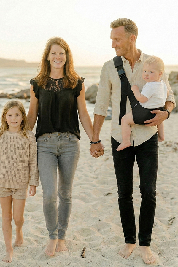 Rodzina idzie boso po plaży o zachodzie słońca; tata niesie malucha w czarnym nosidełku na biodro DereDere z szeroką szelką i klamrą, obok mama i starsza córka.