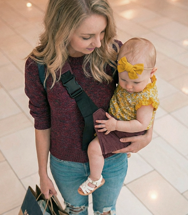 Kobieta w bordowym swetrze i podartych jeansach niesie dziewczynkę w żółtej bluzeczce i opasce. Dziecko siedzi stabilnie w bordowym nosidełku na biodro DereDere z szeroką, czarną szelką; tło galerii handlowej.