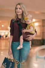 Kobieta w bordowym swetrze i podartych jeansach niesie dziewczynkę w żółtej bluzeczce i opasce. Dziecko siedzi stabilnie w bordowym nosidełku na biodro DereDere z szeroką, czarną szelką; tło galerii handlowej.