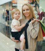 Mama na zakupach trzyma dziecko w czarnym nosidełku na biodro DereDere; czarna szelka z dużą klamrą, torby w ręku, uśmiechy.