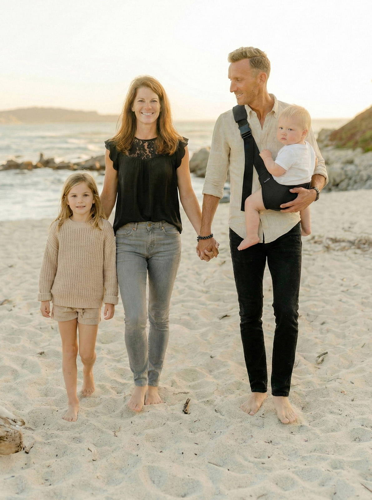 Rodzina idzie boso po plaży; tata ma nosidełko ergonomiczne DereDere i niesie w nim malucha, obok mama i starsza córka, złote światło zachodu.