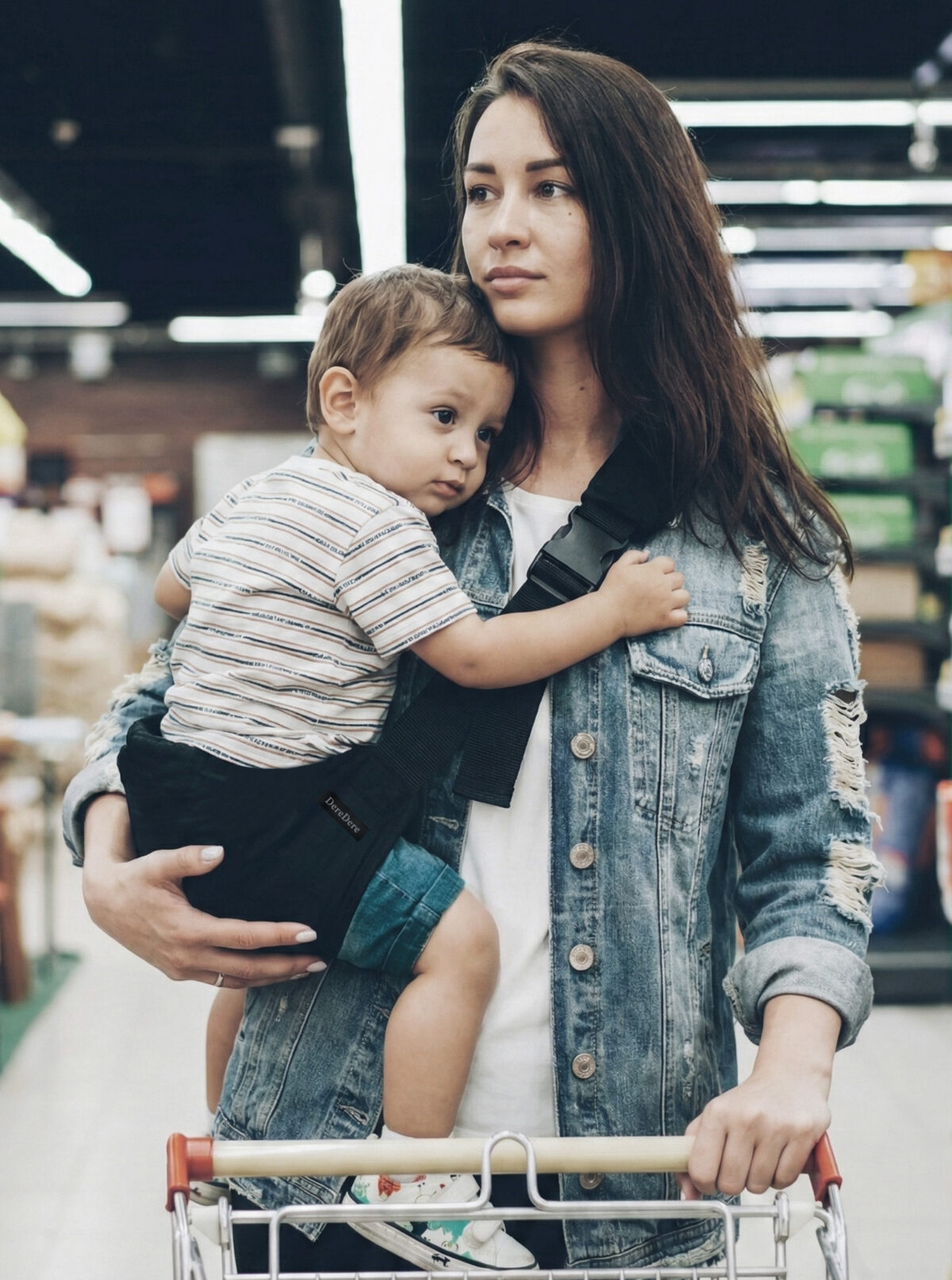 Kobieta w dżinsowej kurtce prowadzi wózek w supermarkecie i niesie synka przytulonego do ramienia. Maluch siedzi stabilnie w czarnym nosidełku ergonomicznym DereDere z szeroką szelką i solidną klamrą.