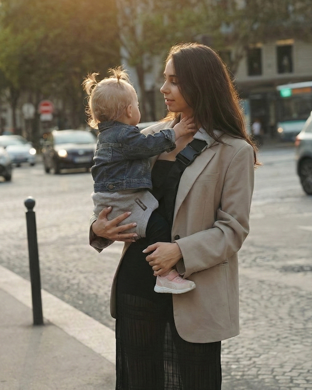Mama na miejskim spacerze trzyma dziecko w szarym nosidełku na biodro DereDere; czarna szelka z klamrą, złota godzina, ruch uliczny w tle.