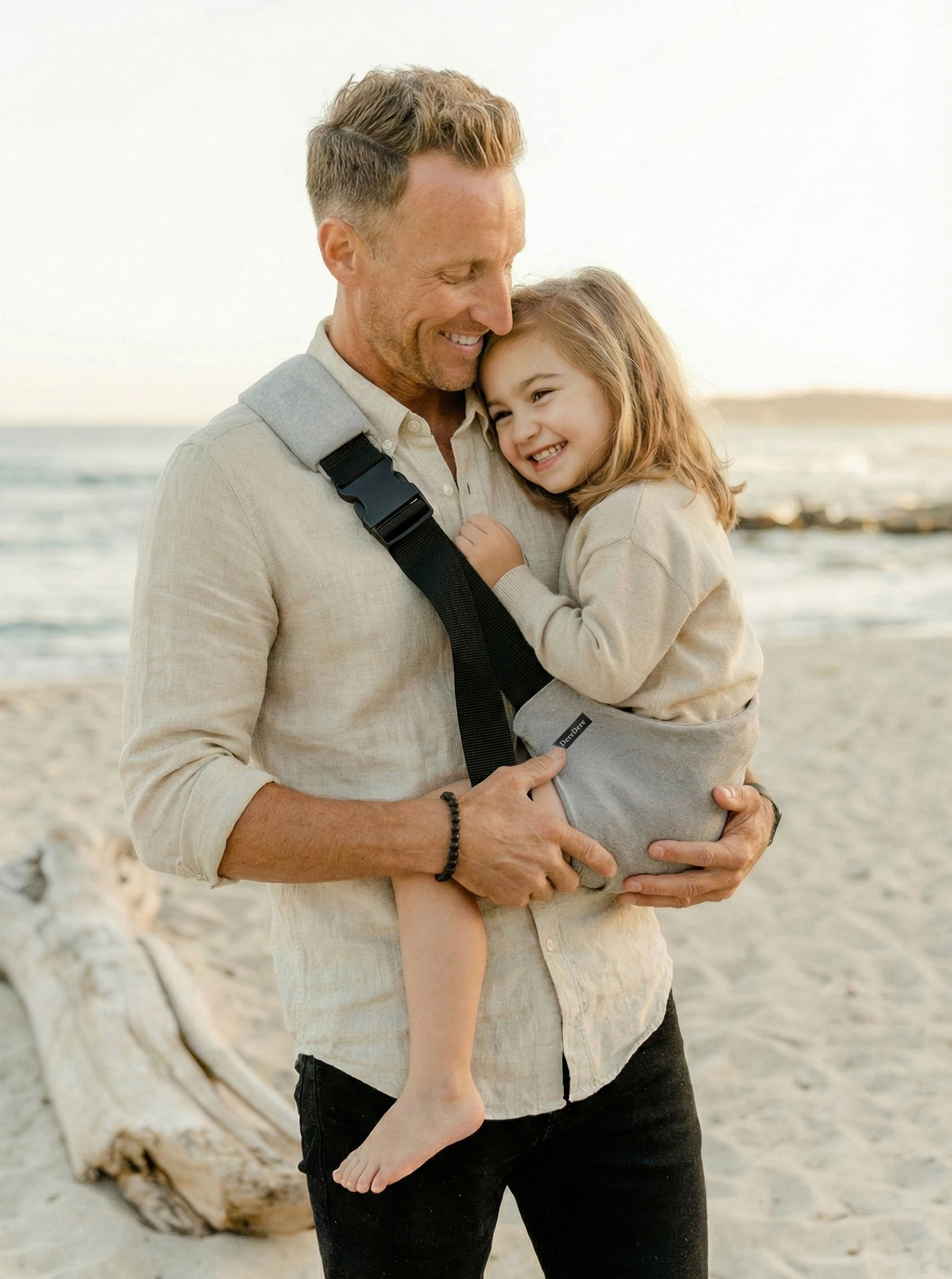 Ciepły kadr z plaży o złotej godzinie. Tata w beżowej, lnianej koszuli i czarnych spodniach ma nosidełko ergonomiczne i obejmuje córkę siedzącą w tym szarym nosidełku. Przez ramię biegnie szeroka czarna szelka z solidną klamrą i miękkim naramiennikiem; rodzic podtrzymuje dziecko dłonią. Dziewczynka wtula się z uśmiechem; w tle piasek, pnie wyrzucone przez morze i spokojna woda.