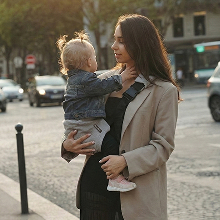 Mama na miejskim spacerze trzyma dziecko w szarym nosidełku na biodro DereDere; szeroka czarna szelka, złote popołudniowe światło.