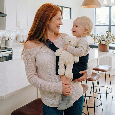 Rudowłosa mama w kuchni trzyma uśmiechnięte dziecko z misiem w czarnym nosidełku dla dzieci DereDere; wygoda w domu i na co dzień.