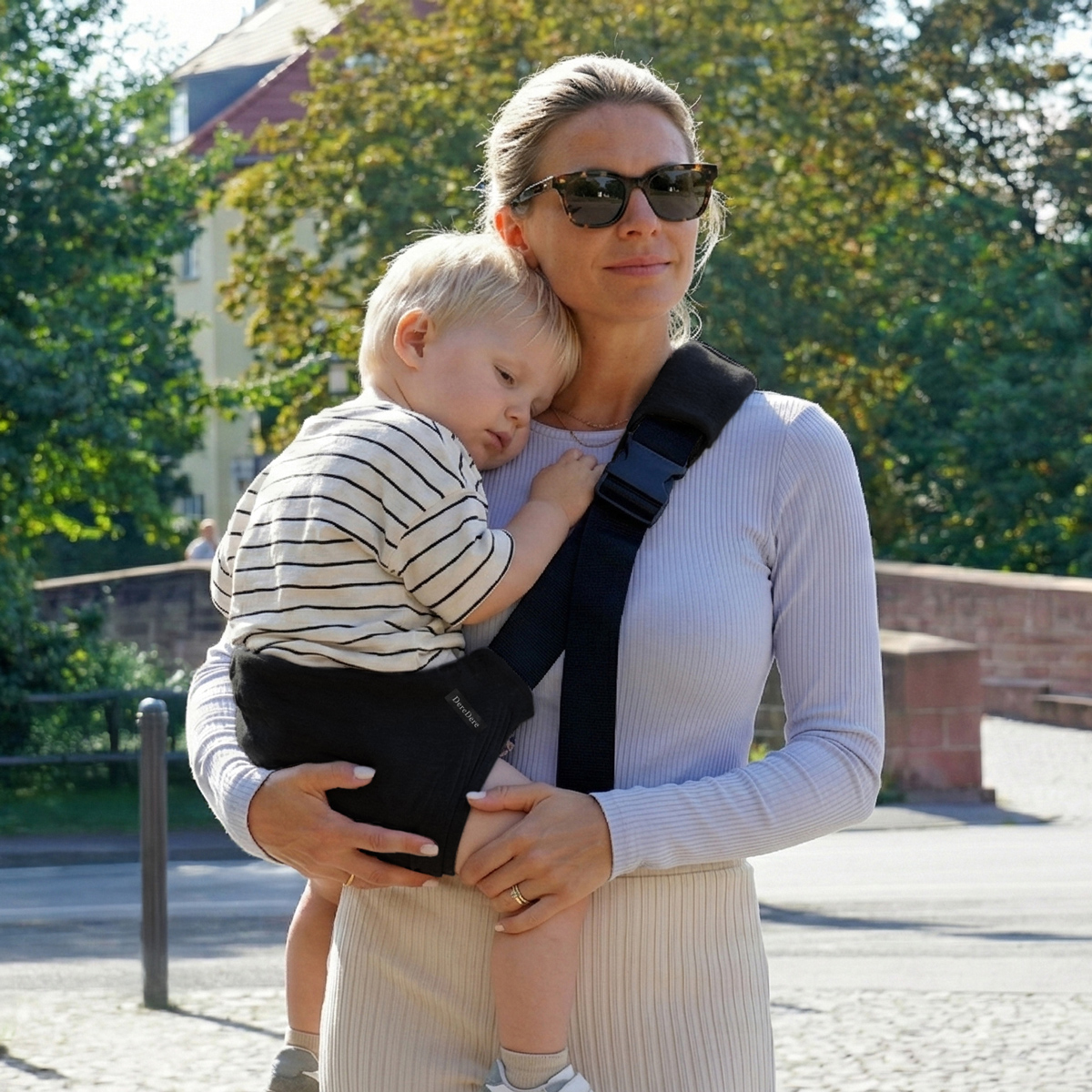 Mama niesie śpiące dziecko w czarnym nosidełku DereDere na biodro podczas spaceru po mieście.