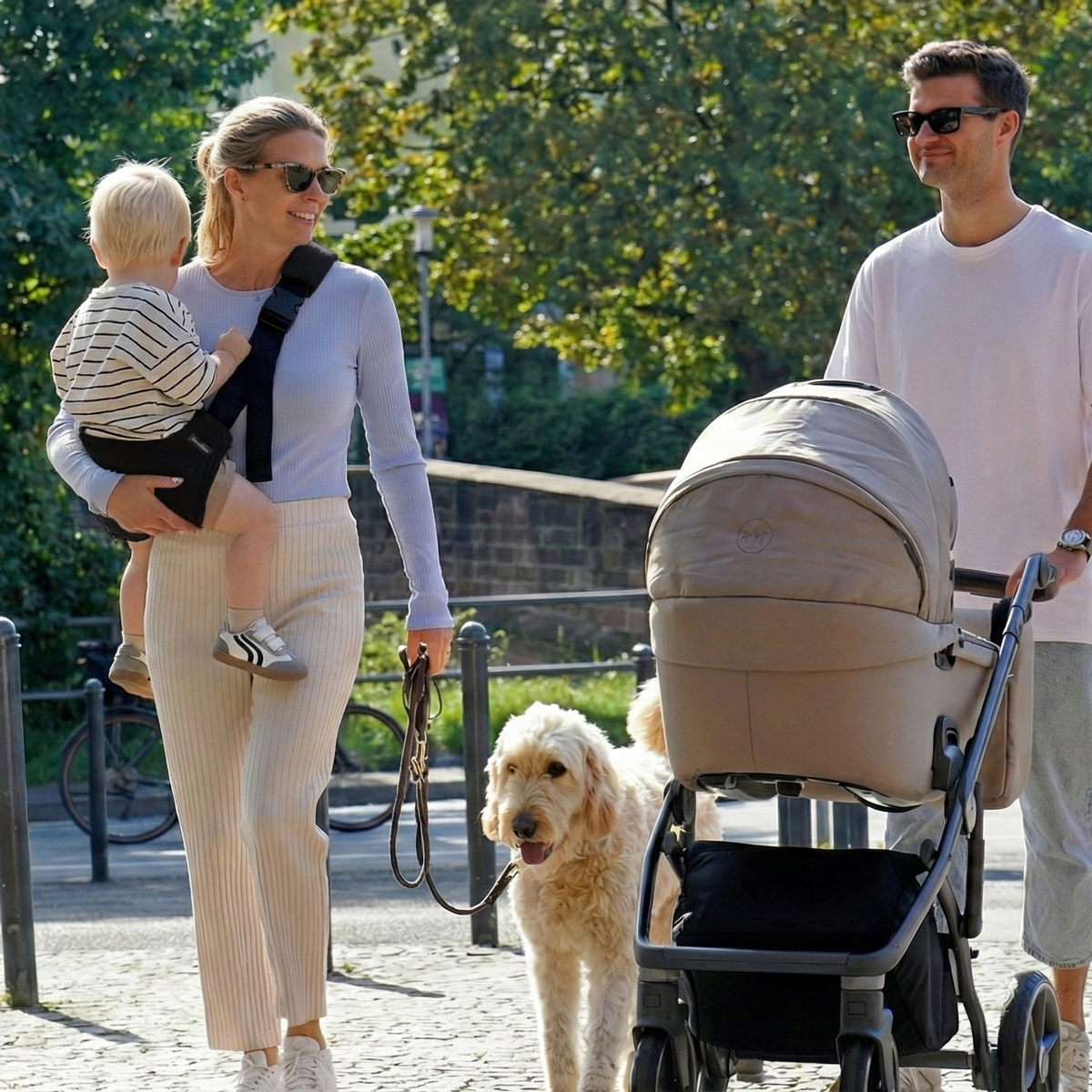 Kobieta na spacerze używa nosidło dla dziecka DereDere do noszenia dziecka, prowadząc psa na smyczy, obok mężczyzna z wózkiem.