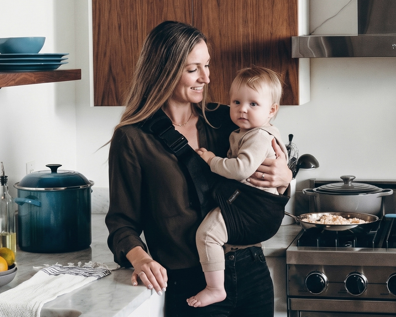 Mama trzyma dziecko w czarnym nosidełku DereDere w kuchni podczas codziennych domowych obowiązków.
