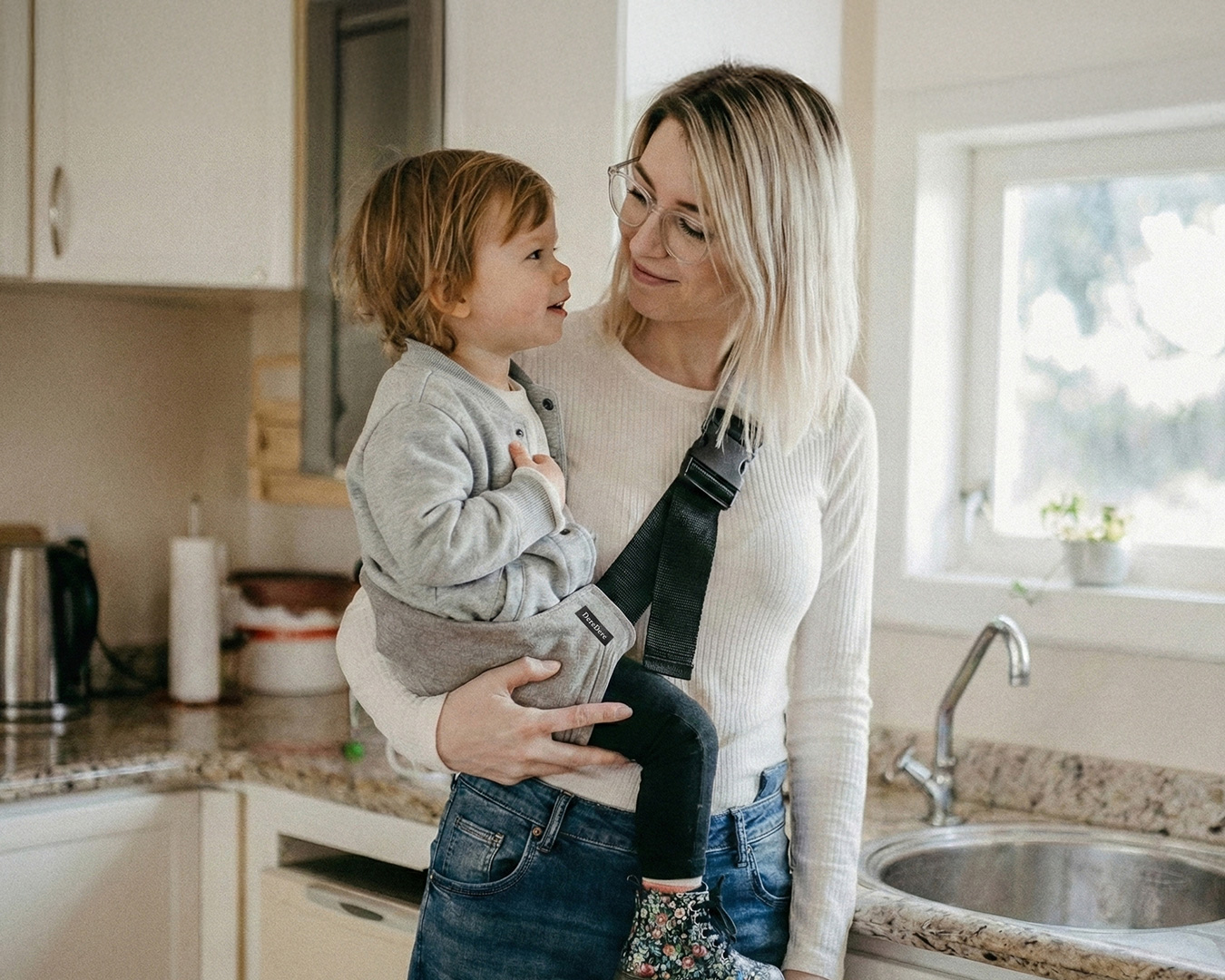 Mama trzyma starsze dziecko w jasnoszarym nosidełku DereDere w kuchni i patrzy na nie z uśmiechem.