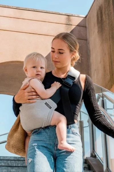 Mama niesie dziecko w jasnoszarym nosidełku dla dziecka DereDere podczas schodzenia po schodach.
