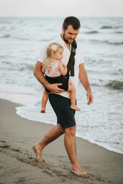 Tata z dzieckiem w czarnym nosidełku dla dziecka DereDere podczas spaceru po plaży.