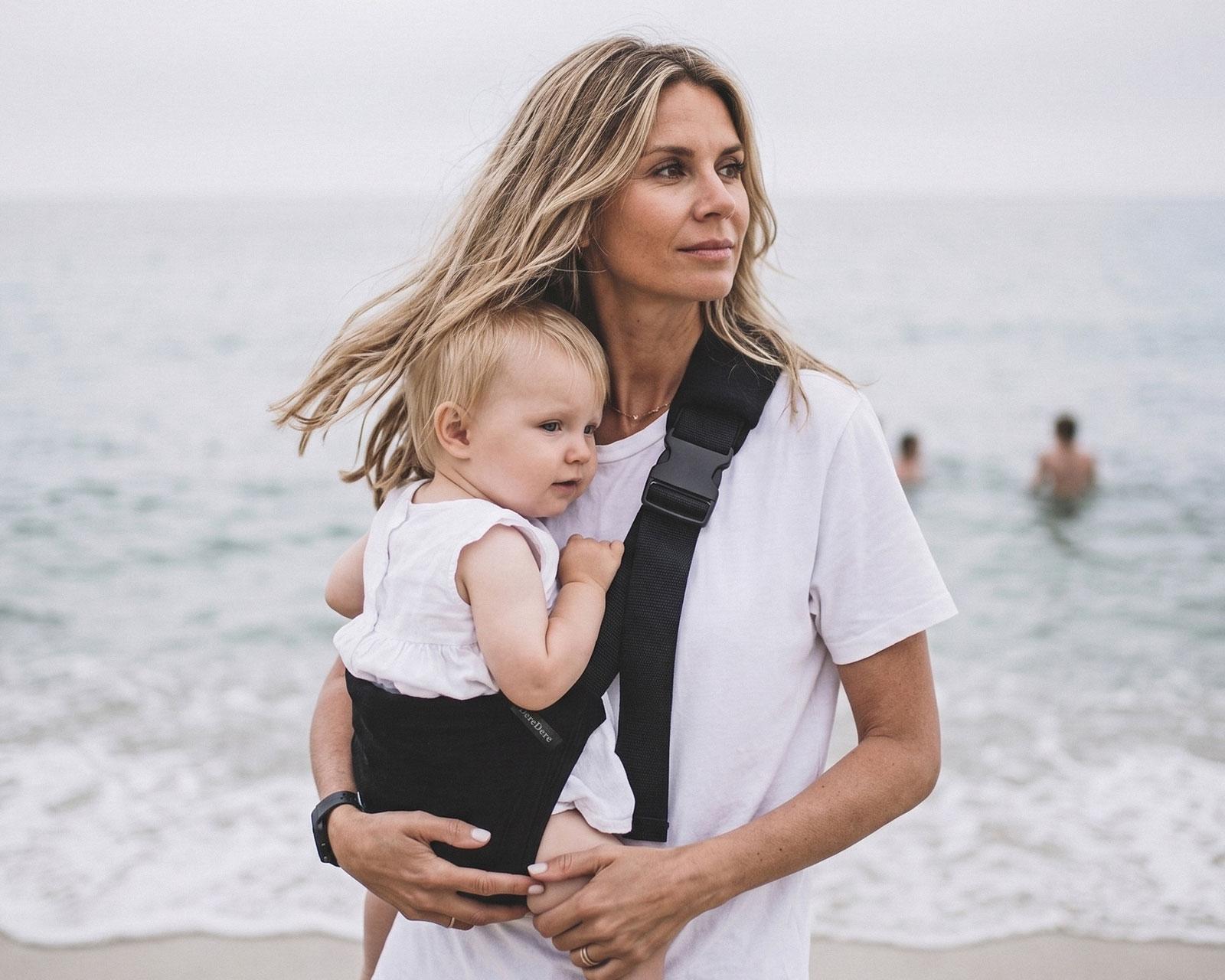 Mama z dzieckiem w czarnym nosidełku dla dziecka DereDere podczas spaceru po plaży.