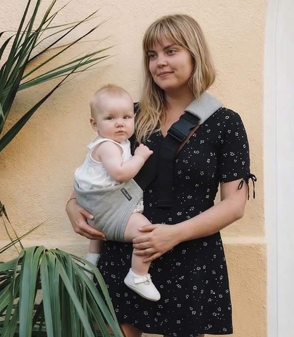 Mama ma szare nosidełko biodrowe w którym nosi dziecko.